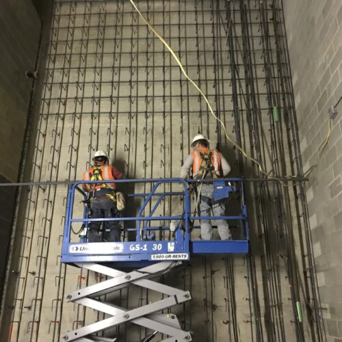 Construction workers on scissor lift repair wall with exposed rebar.