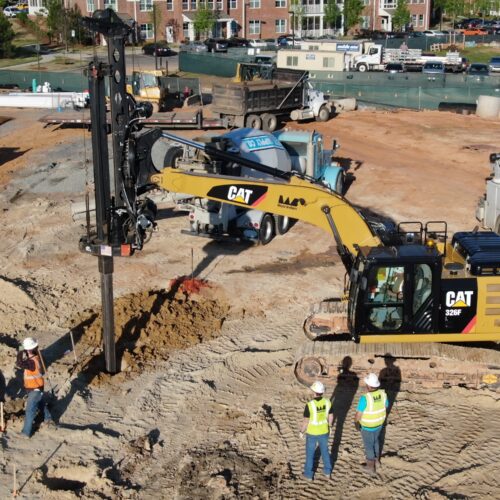 Workers watch CAT excavator drive steel pile at site.