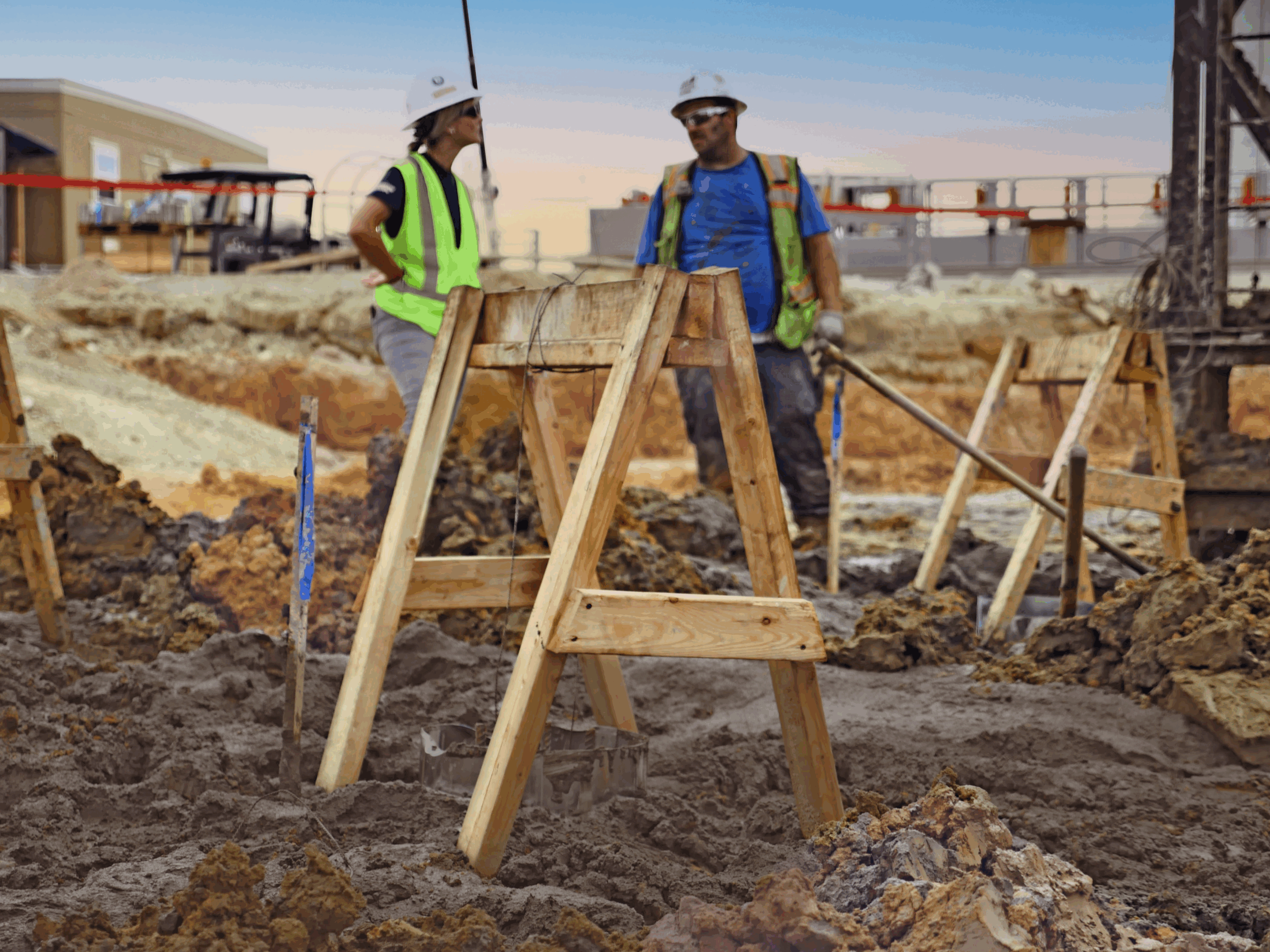 Two construction workers in safety gear at a muddy site.