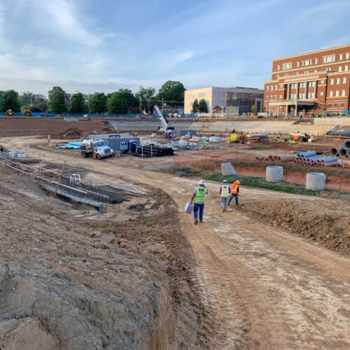 Workers walk on dirt path at busy construction site, brick building behind.