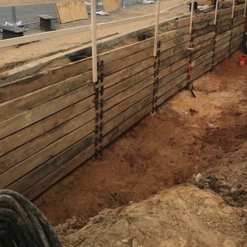 Two workers in vests by trench, wooden wall, red buckets.