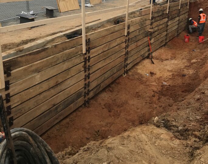 Two workers in vests by trench, wooden wall, red buckets.