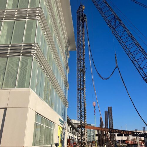Cranes build steel framework near glass building, muddy ground.