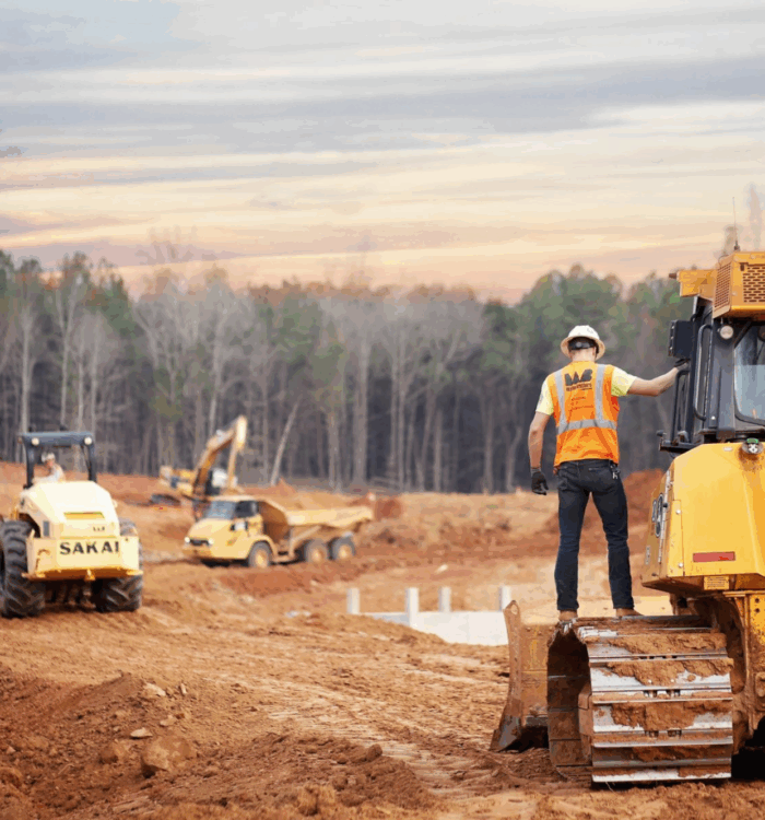 Worker in safety vest stands on bulldozer at dirt site.