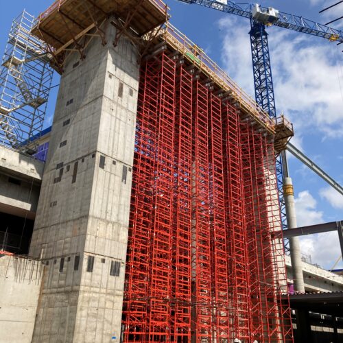 Tall concrete column on red scaffolding, blue crane behind.