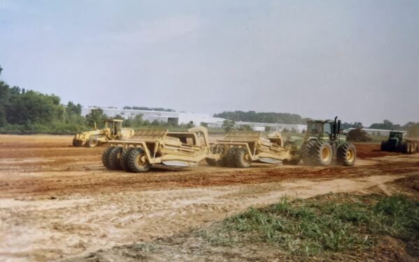 Yellow construction vehicles lined up on dirt, trees behind.