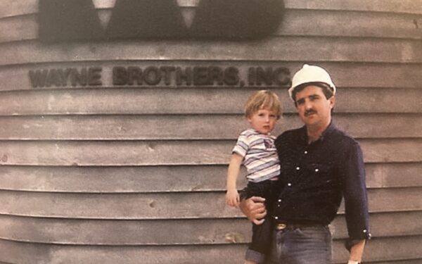 Man in hard hat holds child before Wayne Brothers, Inc. signage