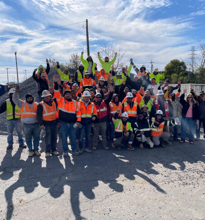 Construction workers in vests and helmets pose together, smiling outdoors.