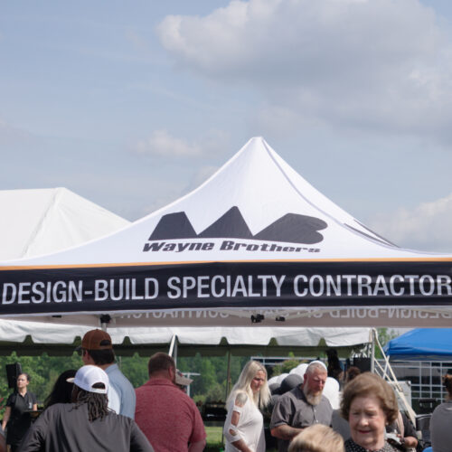 Crowd in front of Wayne Brothers Design-Build tent outdoors.