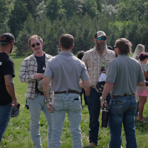 Six men chat with drinks outdoors near balloons and trees.