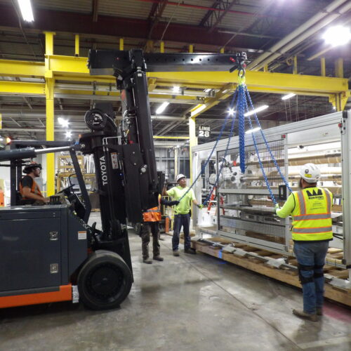 Workers move equipment with forklift, hoist; wearing safety gear.