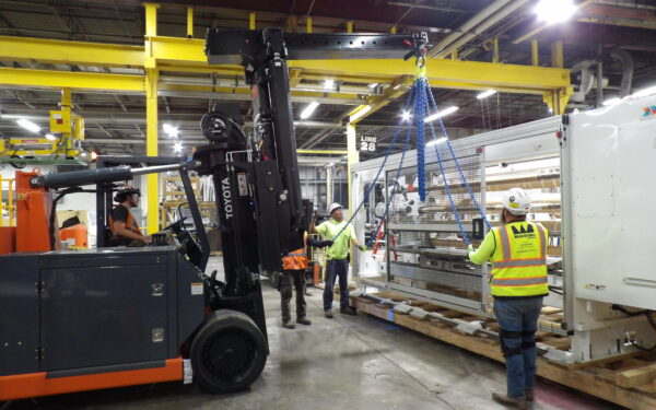 Workers move equipment with forklift, hoist; wearing safety gear.