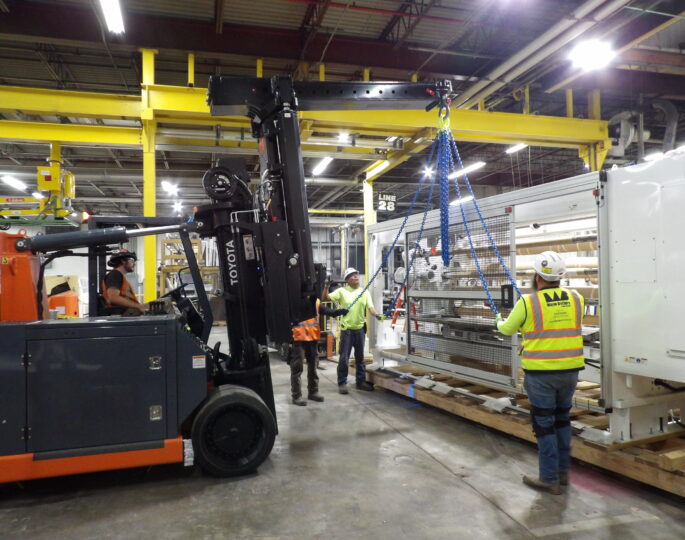 Workers move equipment with forklift, hoist; wearing safety gear.
