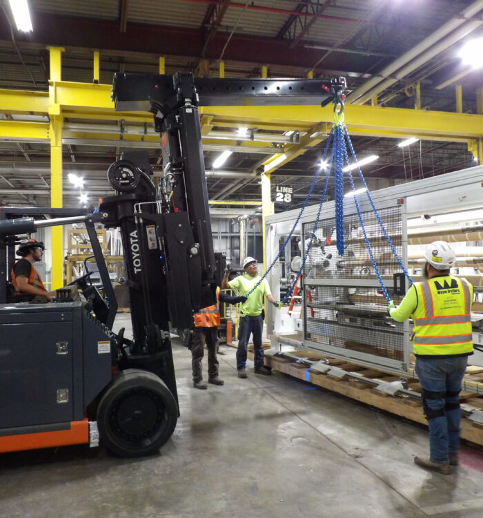 Workers move equipment with forklift, hoist; wearing safety gear.