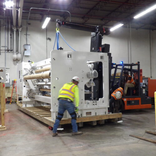 Two workers in safety gear move a large machine with forklifts.