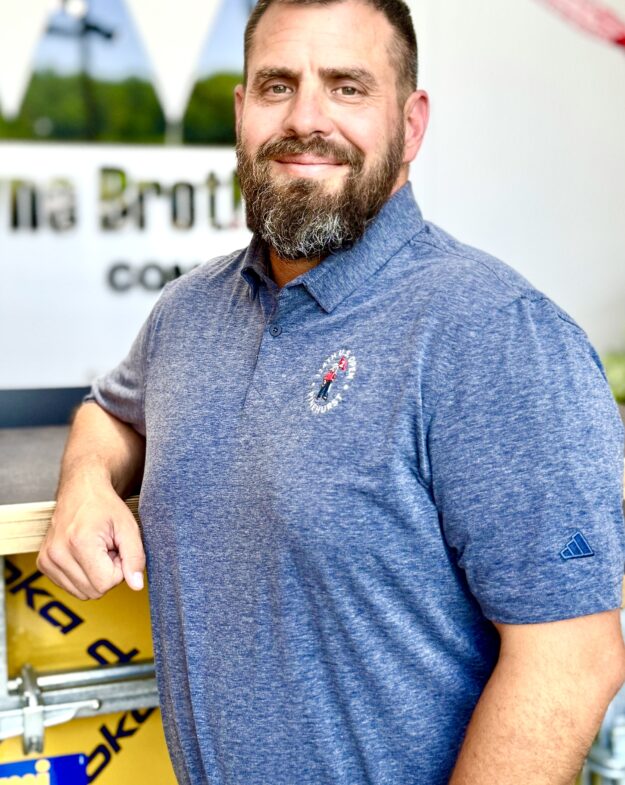 Bearded man in blue polo leans on yellow barrier indoors.