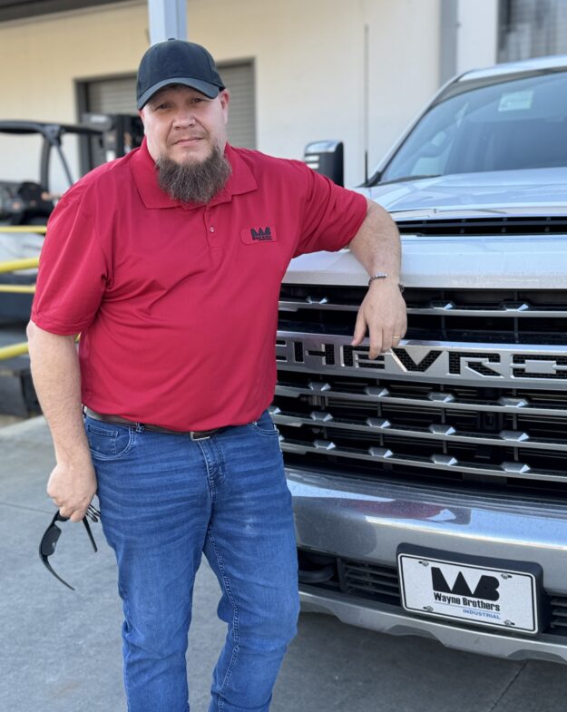 Man in red polo leans on silver truck outside building.