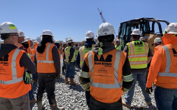 Construction workers in safety gear stand by equipment on gravel.
