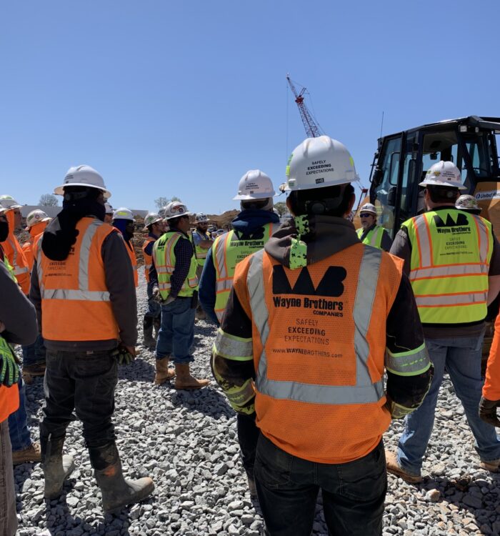 Construction workers in safety gear stand by equipment on gravel.