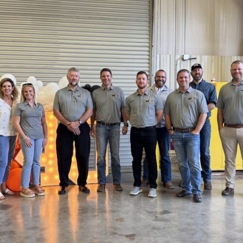 Eleven people in gray shirts pose in a warehouse.