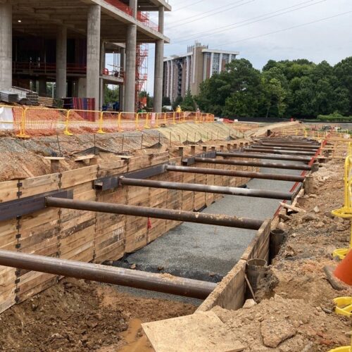 Construction site: wooden retaining walls, steel beams, workers in gear.