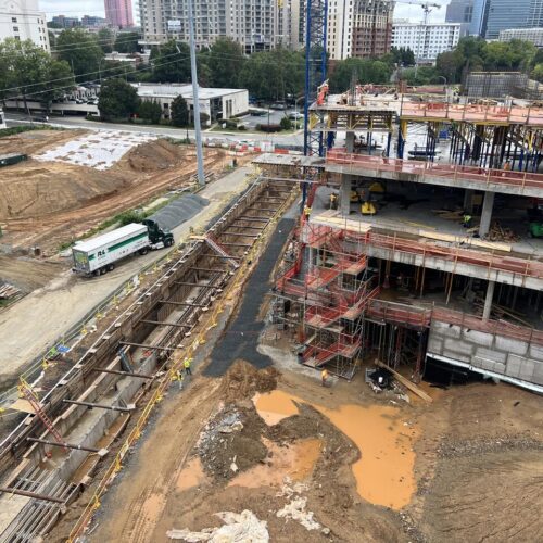 Partially built high-rise, steel beams, machinery, mud, city backdrop.
