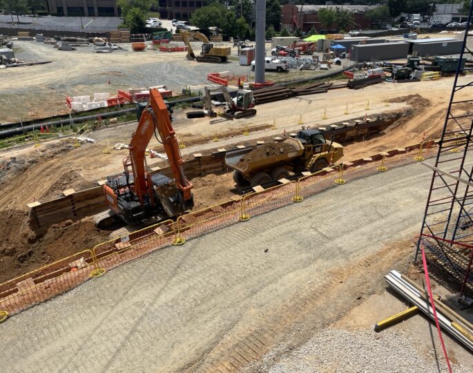Excavator and dump truck working in a sunny, barriered trench.
