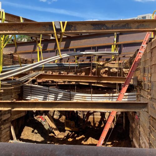 Beams support excavation site with pipes, ladder, materials visible.