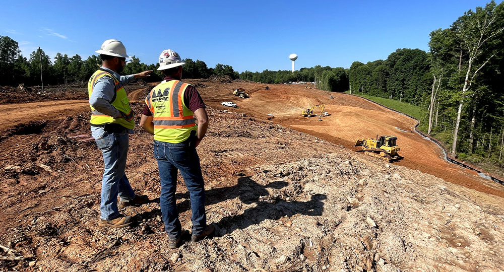 Two workers in safety gear watch machinery on sunny construction site.