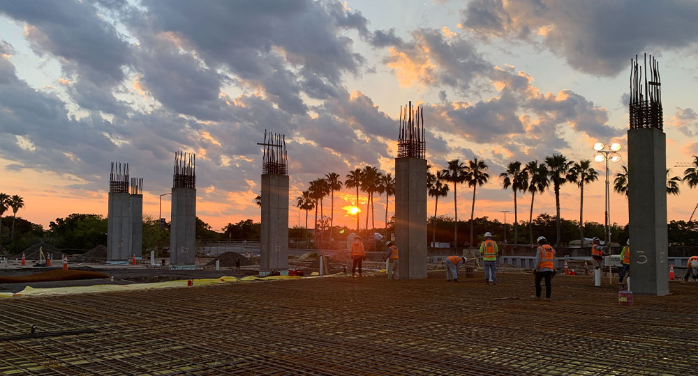 Workers build at sunset; columns, rebar, and palm trees visible.