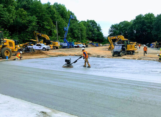 Workers and machines pave concrete amid surrounding trees.