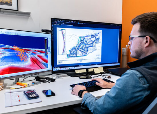 Man at desk with two monitors displaying 3D models.
