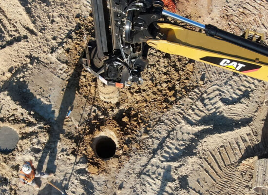 CAT excavator drills; worker stands on dirt nearby.