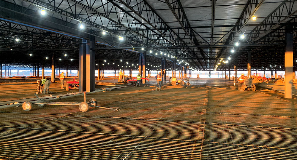Workers install steel rebar under lights in open indoor site.