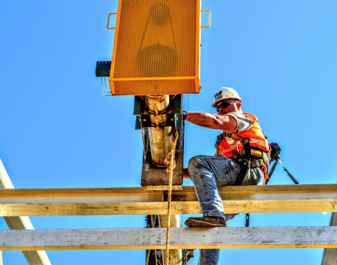 Worker in safety gear operates equipment on steel beam, blue sky.