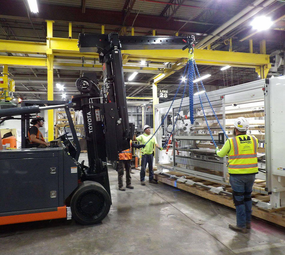 Workers use forklift and straps to move machinery in warehouse.