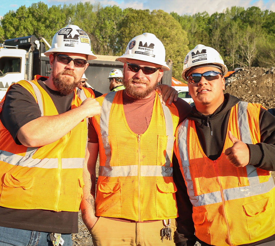 Three construction workers pose together outdoors, wearing safety gear.