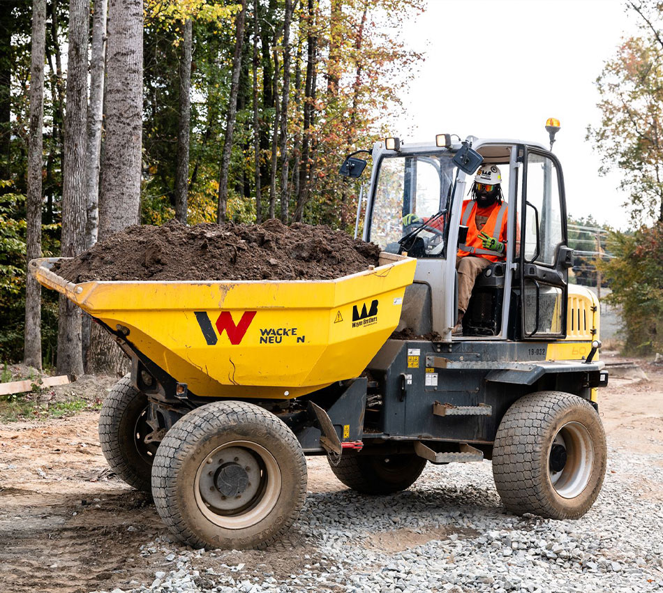 Worker drives yellow dump truck of soil on gravel path.