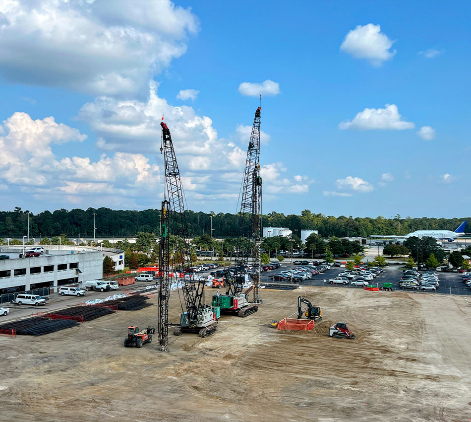 Two cranes, construction vehicles on dirt lot by parking area.