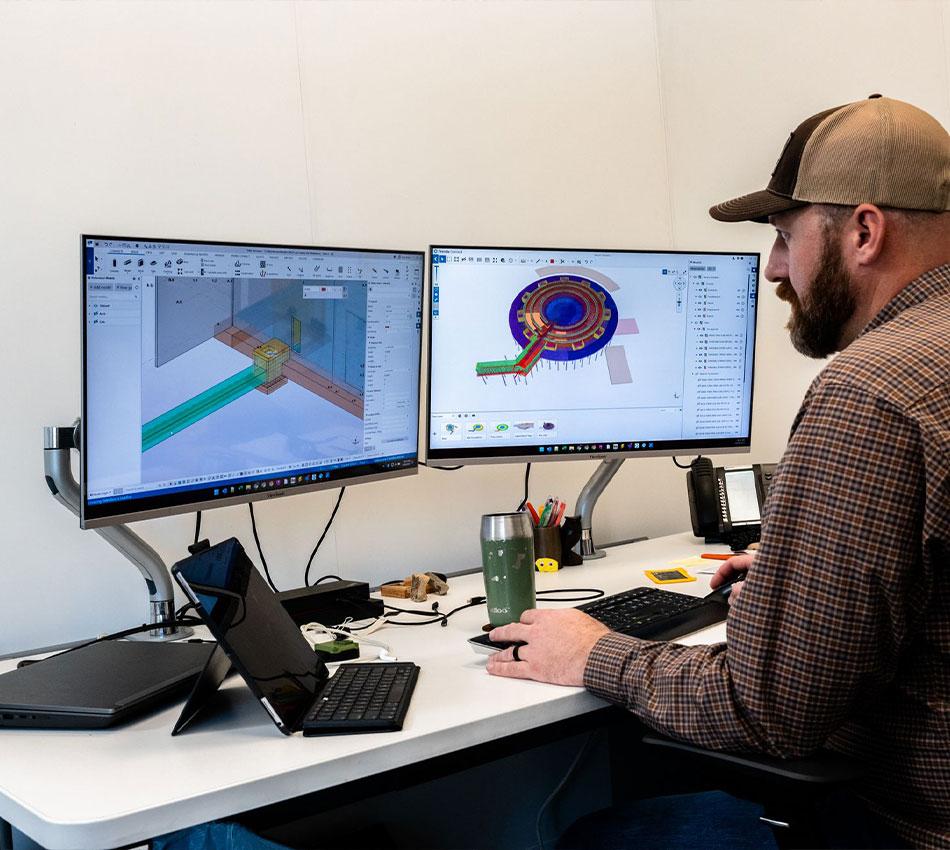 Man at desk with two monitors, 3D models, laptop, tablet.