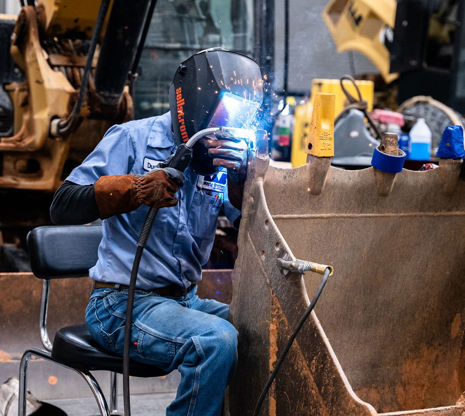 Worker in protective gear welds metal while seated in workshop.