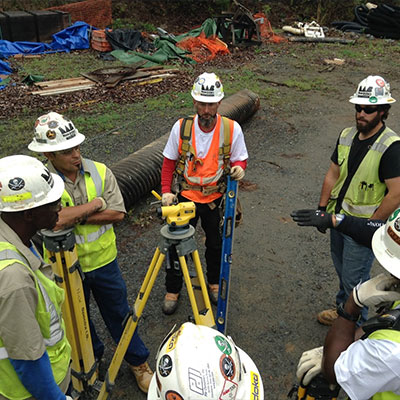 Construction workers in safety gear discuss a task around equipment.