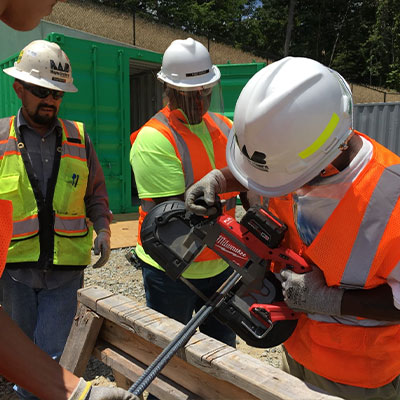 Three workers cut metal rebar at worksite with containers.
