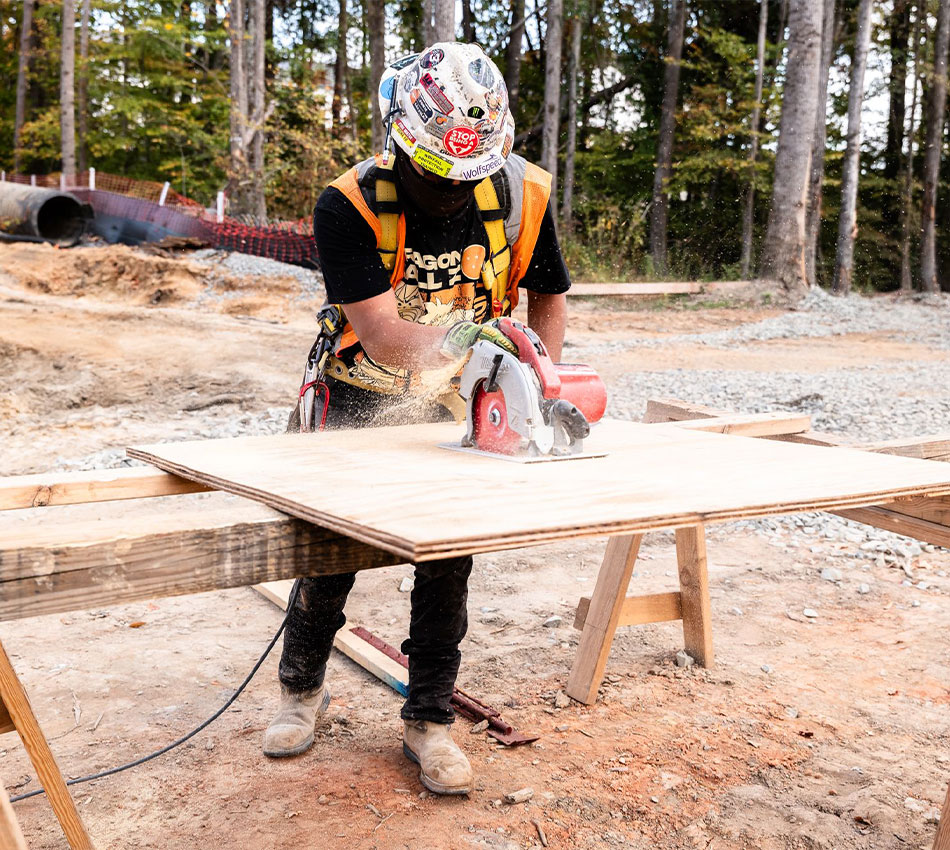 Worker in safety gear cuts plywood with saw at outdoor site.