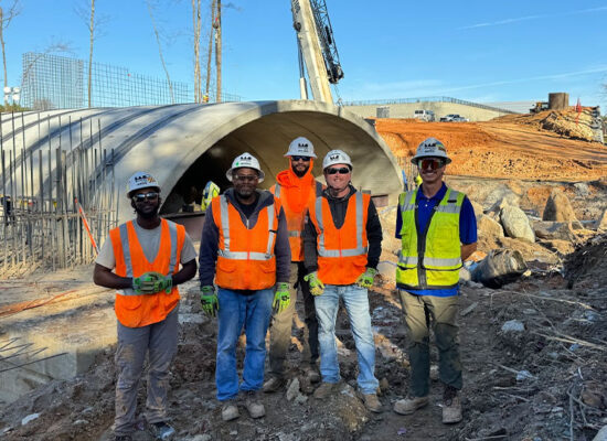 Five workers in safety gear stand by unfinished concrete structure.