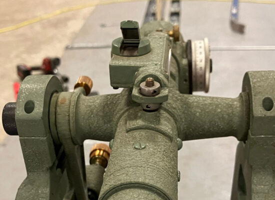 Green theodolite with knobs and scope on indoor tripod.