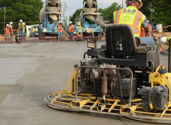 Construction worker smooths concrete with ride-on trowel; others nearby.