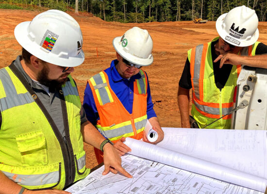 Three workers in safety gear examine blueprints at worksite.