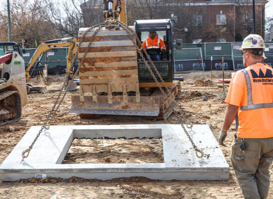 Workers use machinery to lift concrete slab with square opening.