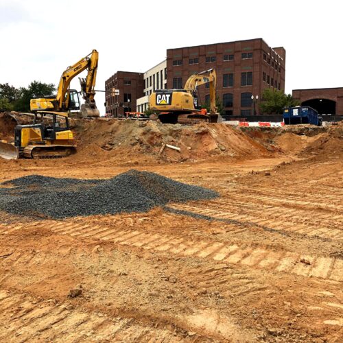 Excavators work on dirt near brick building and gravel pile.